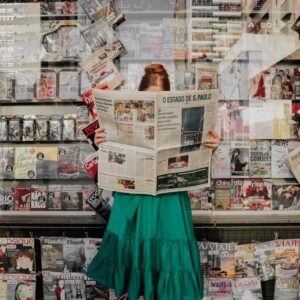 Woman in green dress reading newspaper at street magazine stand outdoors.
