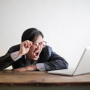 Modern Asian man in jacket and glasses looking at laptop and screaming with mouth wide opened on white background