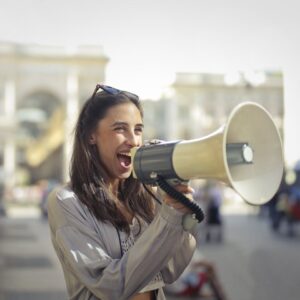 Cheerful young woman in a casual outfit shouting into a megaphone on a sunny day.