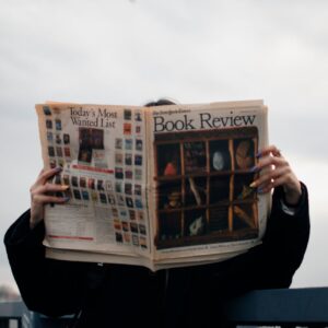 Individual holding a book review newspaper with colorful nails against a cloudy sky backdrop.