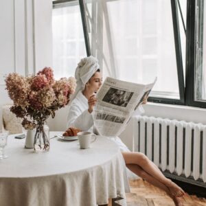 Woman in bathrobe reading newspaper by window, enjoying breakfast with coffee and croissant.