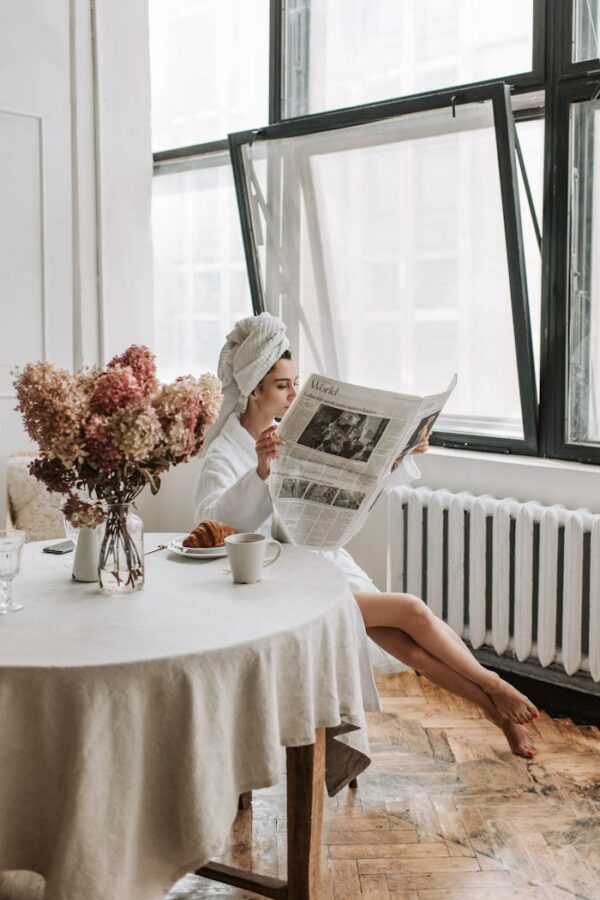 Woman in bathrobe reading newspaper by window, enjoying breakfast with coffee and croissant.